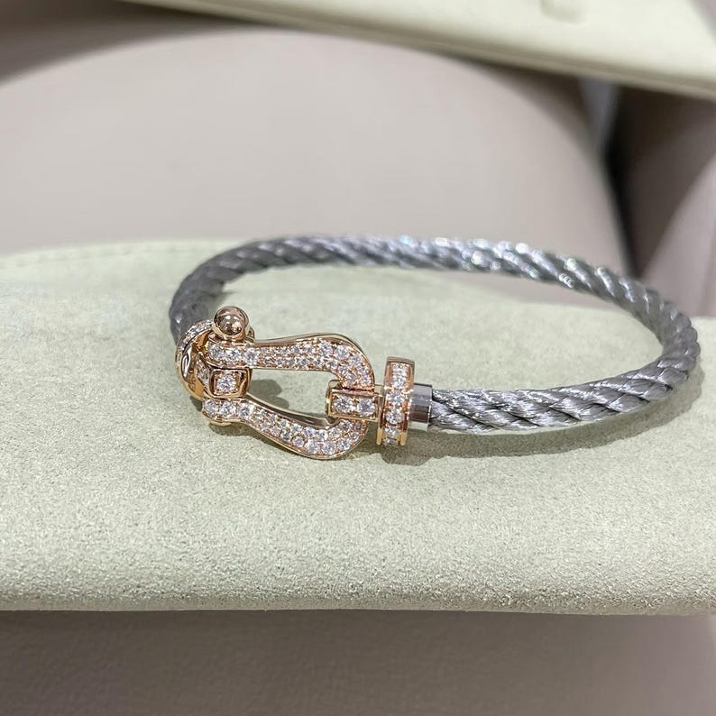 May include: A silver cable bracelet with a rose gold clasp. The clasp, shaped like a shackle, is set with small, clear gemstones. The bracelet is presented on a light green surface.