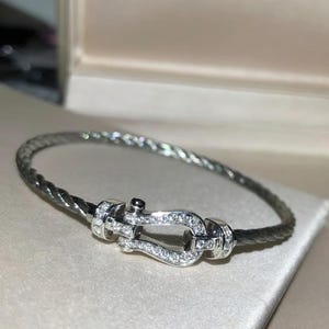 May include: A silver bracelet with a braided band and a decorative clasp. The clasp is adorned with small, sparkling gemstones. The bracelet is displayed on a light-colored surface, suggesting a jewelry theme.