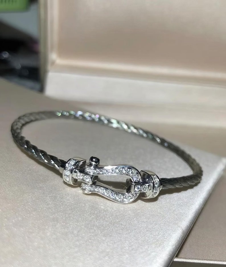May include: A silver bracelet with a braided band and a decorative clasp. The clasp is adorned with small, sparkling gemstones. The bracelet is displayed on a light-colored surface, suggesting a jewelry theme.