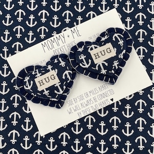 Pu&ograve; includere: Due abbracci tascabili a forma di cuore in feltro con la parola "HUG" su ciascuno. I cuori sono blu navy con un motivo di ancora bianca e cuciture bianche. I cuori sono su una carta bianca con il testo "Mommy + Me Pocket Hugs" e "Fianco a fianco o a miglia di distanza, saremo sempre connessi da questi due cuori".