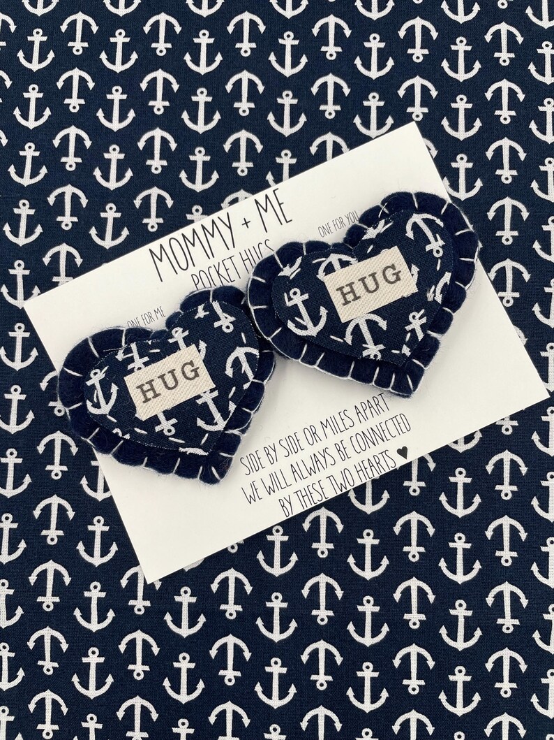 Pu&ograve; includere: Due abbracci tascabili a forma di cuore in feltro con la parola "HUG" su ciascuno. I cuori sono blu navy con un motivo di ancora bianca e cuciture bianche. I cuori sono su una carta bianca con il testo "Mommy + Me Pocket Hugs" e "Fianco a fianco o a miglia di distanza, saremo sempre connessi da questi due cuori".