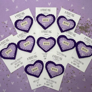 May include: Nine purple felt heart-shaped pouches with the word "HUG" printed on them. Each pouch is attached to a white card with the text "A Pocket Hug" printed on it.