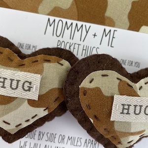 Pu&ograve; includere: Due cuori di feltro marroni a forma di tasca con un motivo mimetico e la parola "HUG" su ciascuno. I cuori sono su una carta bianca con il testo "Mommy + Me Pocket Hugs" e "One for me, One for you".