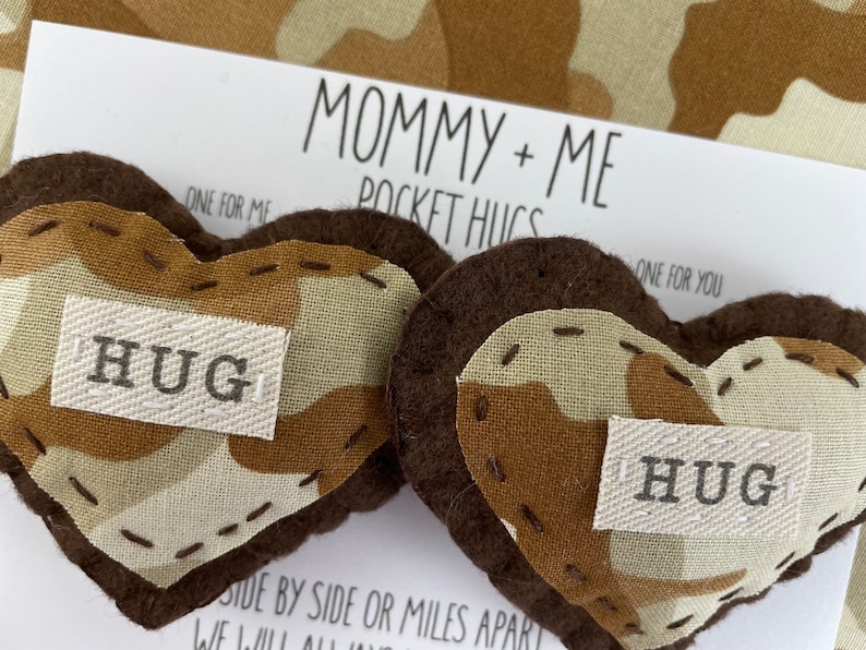 Pu&ograve; includere: Due cuori di feltro marroni a forma di tasca con un motivo mimetico e la parola "HUG" su ciascuno. I cuori sono su una carta bianca con il testo "Mommy + Me Pocket Hugs" e "One for me, One for you".