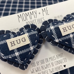 May include: Two felt heart-shaped pocket hugs with a blue and white plaid pattern. Each heart has a white tag with the word "HUG" printed on it. The hearts are attached to a white card with the text "Mommy + Me Pocket Hugs" and "Side by side or miles apart we will always be connected by these two hearts".