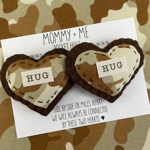 Pu&ograve; includere: Due abbracci tascabili a forma di cuore in feltro marrone con un motivo mimetico e la parola "HUG" su ciascuno. I cuori sono su una carta bianca con il testo "Mommy + Me Pocket Hugs" e "Fianco a fianco o a miglia di distanza, saremo sempre connessi da questi due cuori".