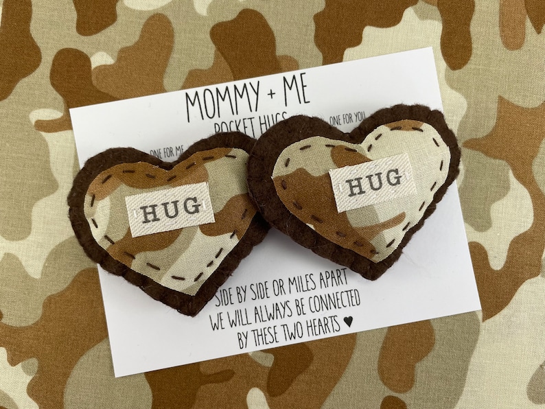 Pu&ograve; includere: Due abbracci tascabili a forma di cuore in feltro marrone con un motivo mimetico e la parola "HUG" su ciascuno. I cuori sono su una carta bianca con il testo "Mommy + Me Pocket Hugs" e "Fianco a fianco o a miglia di distanza, saremo sempre connessi da questi due cuori".