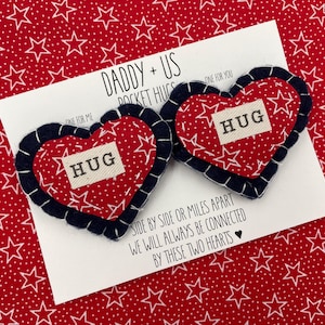 Pu&ograve; includere: Due abbracci tascabili a forma di cuore in feltro rosso con stelle bianche e la parola "HUG" su ciascuno. I cuori sono cuciti con un bordo blu scuro. I cuori sono su una carta bianca con il testo "Daddy + Us Pocket Hugs" e "Side by side or miles apart we will always be connected by these two hearts".
