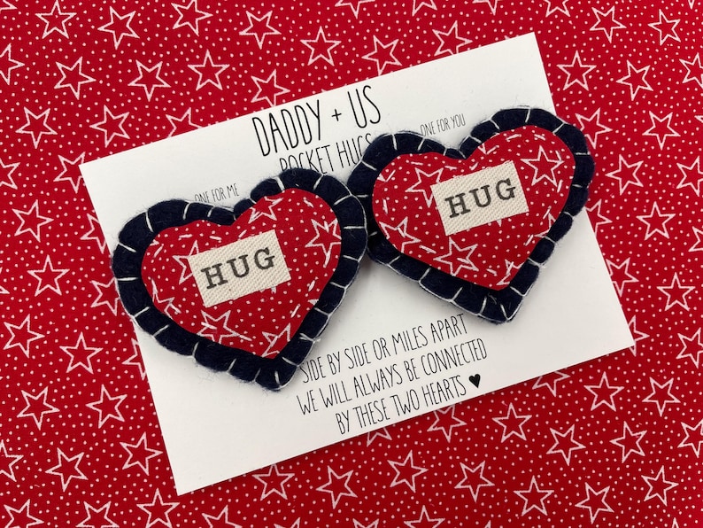 Pu&ograve; includere: Due abbracci tascabili a forma di cuore in feltro rosso con stelle bianche e la parola "HUG" su ciascuno. I cuori sono cuciti con un bordo blu scuro. I cuori sono su una carta bianca con il testo "Daddy + Us Pocket Hugs" e "Side by side or miles apart we will always be connected by these two hearts".