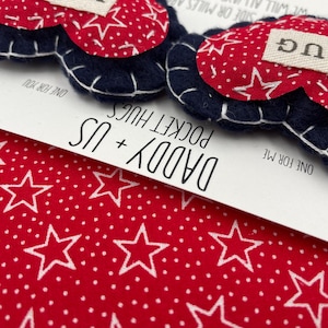 Pu&ograve; includere: Due abbracci tascabili a forma di cuore in feltro con motivi a stelle rosse e bianche e bordi blu. Un cuore ha la parola "HUG" su di esso. I cuori sono su una carta bianca con il testo "Daddy + Us Pocket Hugs" e "One for me" e "One for you".