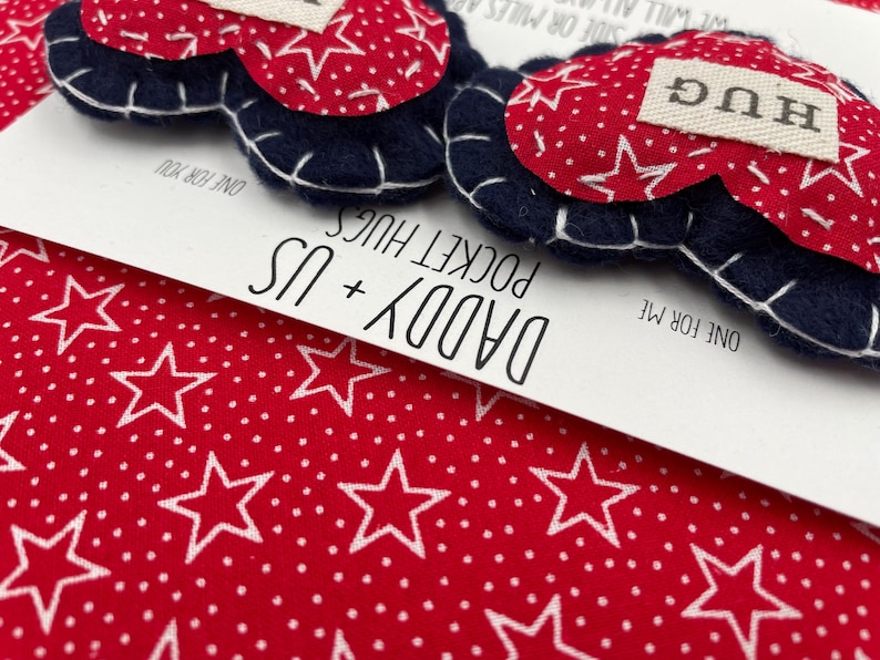 Pu&ograve; includere: Due abbracci tascabili a forma di cuore in feltro con motivi a stelle rosse e bianche e bordi blu. Un cuore ha la parola "HUG" su di esso. I cuori sono su una carta bianca con il testo "Daddy + Us Pocket Hugs" e "One for me" e "One for you".
