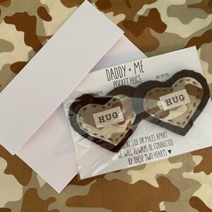 Pu&ograve; includere: Due abbracci tascabili a forma di cuore in feltro con la parola "HUG" ricamata su ciascuno. I cuori sono marroni con un motivo mimetico beige. Il testo "Daddy + Me Pocket Hugs" e "One for you" &egrave; stampato su un cartoncino bianco.