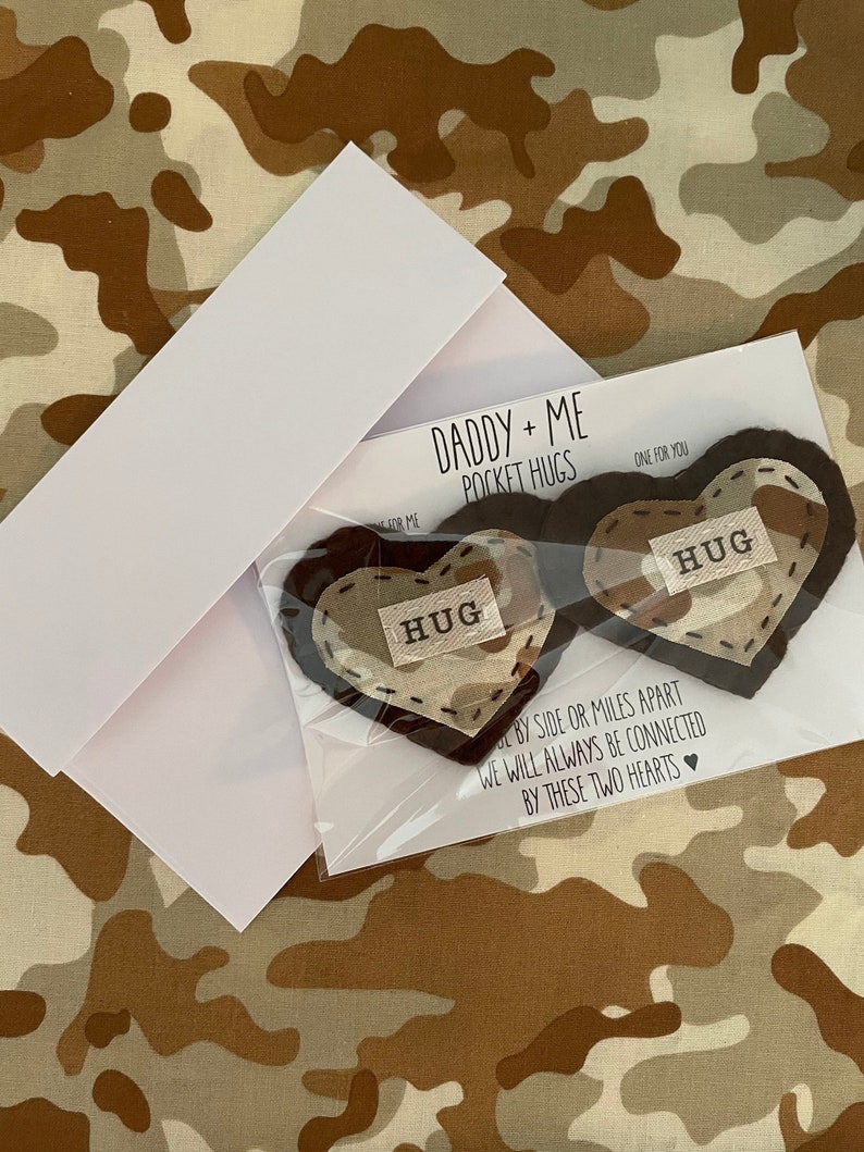 Pu&ograve; includere: Due abbracci tascabili a forma di cuore in feltro con la parola "HUG" ricamata su ciascuno. I cuori sono marroni con un motivo mimetico beige. Il testo "Daddy + Me Pocket Hugs" e "One for you" &egrave; stampato su un cartoncino bianco.
