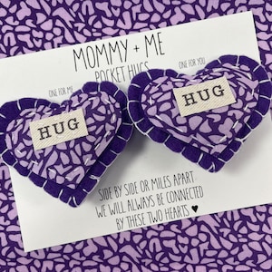 Peut inclure: Deux cœurs en feutre violet en forme de cœur avec le mot "HUG" sur chacun. Les cœurs sont sur une carte blanche avec le texte "Mommy + Me Pocket Hugs" et "Cote à cote ou à des kilomètres de distance, nous serons toujours connectés par ces deux cœurs".