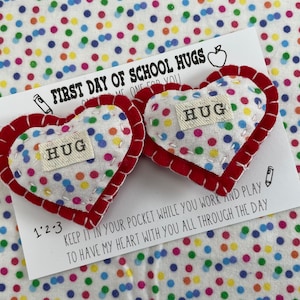 May include: Two small felt hearts with red trim and colorful polka dots. Each heart has a white tag sewn on with the word "HUG" printed on it. The hearts are on a white card with the text "FIRST DAY OF SCHOOL HUGS" and "KEEP IT IN YOUR POCKET WHILE YOU WORK AND PLAY TO HAVE MY HEART WITH YOU ALL THROUGH THE DAY".