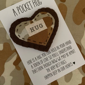 Può includere: Un cuore di feltro marrone con un motivo mimetico beige e marrone. Il cuore ha un'etichetta bianca cucita su di esso che dice "HUG". Il cuore è su una carta bianca con il testo "A Pocket Hug" e un messaggio sugli abbracci.