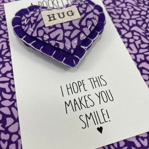 May include: A purple felt heart with the word "HUG" stitched on it. The heart is attached to a white card with the text "I hope this makes you smile!"