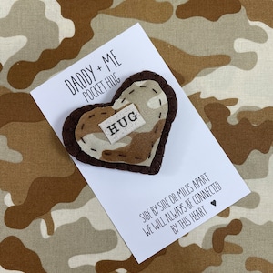 May include: A heart-shaped pocket hug with a camouflage pattern and the word "HUG" on a small tag. The card reads "DADDY + ME POCKET HUG" and "SIDE BY SIDE OR MILES APART WE WILL ALWAYS BE CONNECTED BY THIS HEART."