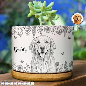 Puede incluir: Una maceta cilíndrica de cerámica blanca con una ilustración de línea negra de un perro Golden Retriever y el nombre Buddy impreso en cursiva negra, decorada con motivos florales, mariposas y una base de madera clara.