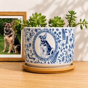 May include: A white ceramic planter with a blue floral pattern and a central oval frame containing a German Shepherd illustration. The planter holds a green succulent plant and sits on a circular wooden saucer, accompanied by a framed photo.
