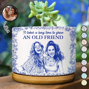 Op de afbeelding: Een witte cilindervormige keramische plantenpot met een houten voet, voorzien van een blauwe lijntekening van twee personen en de tekst "It takes a long time to grow AN OLD FRIEND." Het ontwerp is omlijst door blauwe bloemmotieven aan de zijkanten.
