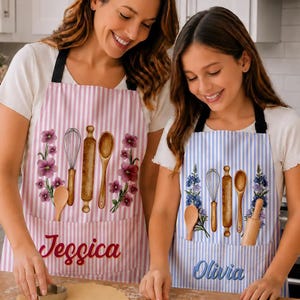 Può includere: Due grembiuli a righe verticali con grafiche di utensili da cucina tra cui una frusta, un mattarello e un cucchiaio di legno. Il grembiule a sinistra è rosa con il nome "Jessica" e quello a destra è blu con il nome "Olivia" in corsivo.
