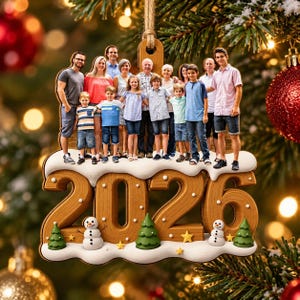 May include: A wooden Christmas ornament featuring a group photo of fourteen people standing on top of large, three-dimensional numbers reading 2026. The base is decorated with white snow, two small snowmen, three green pine trees, and yellow stars.