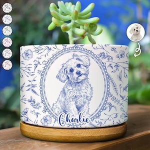 Op de afbeelding: Een cilindervormige plantenbak van wit keramiek met een blauwe lijntekening van een kleine hond in een ovaal kader, met de naam Charlie eronder geschreven. De plantenbak is versierd met blauwe botanische patronen en staat op een ronde houten voet.