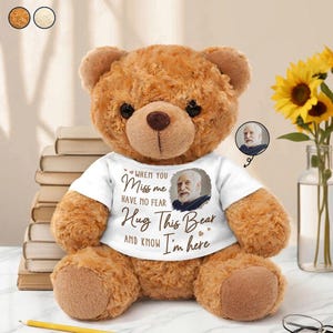 May include: A brown plush teddy bear wearing a white t-shirt printed with the text "WHEN YOU MISS ME HAVE NO FEAR HUG THIS BEAR AND KNOW I'M HERE" and a circular portrait of an older man with a white beard, featuring dark eyes and a dark nose.