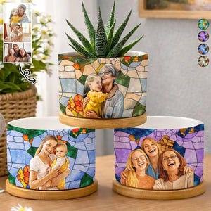May include: Three cylindrical ceramic planters with wooden bases, each featuring a colorful stained-glass style print of family portraits. The top planter holds a green succulent, while the others display illustrations of people.