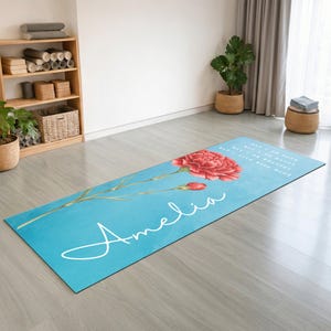 May include: A rectangular yoga mat with a bright blue background featuring a large red carnation flower and the name Amelia written in white cursive script. The mat includes faint white text printed near the top edge of the design.