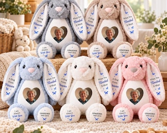 May include: Five plush bunny rabbits in grey, tan, blue, white, and pink. Each features a heart-shaped photo frame on the chest, long ears with the text 'A Hug From Heaven' and 'When You Needed It Most', and feet labelled 'I Am Always With You' and 'GRANDMA'.