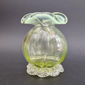 Puede incluir: Un jarrón de cristal transparente con un cuerpo bulboso y una parte superior con volantes y acampanada. El jarrón tiene un tinte verde claro en la base y la parte superior, con un centro transparente. La base tiene un borde festoneado. El jarrón probablemente se utiliza con fines decorativos.