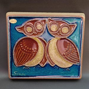 Może przedstawiać: Kwadratowa płytka ceramiczna z dwoma stylizowanymi sowami. Sowy mają brązowe ciała, złote skrzydła i duże, okrągłe oczy. Tło jest turkusowe z błyszczącym wykończeniem. Płytka jest podpisana "Simon".