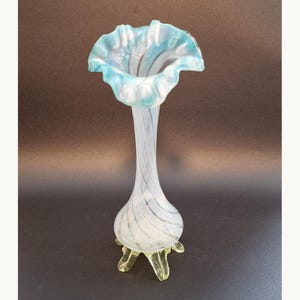 Puede incluir: Un elegante jarrón de cristal con borde acampanado y ondulado en tonos azules y blancos. El jarrón tiene un cuerpo delgado y en espiral y descansa sobre tres pies amarillo-verdosos. Una pieza decorativa para arreglos florales, de unos 25 cm de altura.
