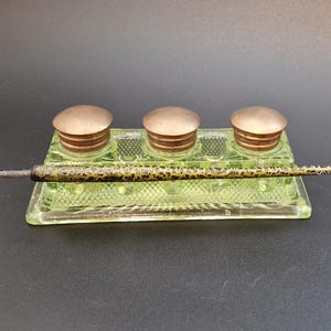 Könnte beinhalten: Ein antikes Tintenfass-Set mit drei Tintenfässern, jedes mit einem Bronzedeckel. Das Tintenfass besteht aus hellgrünem Glas und weist ein dekoratives Muster auf. Ein Stift mit gemustertem Design liegt auf dem Tintenfass.