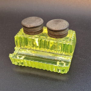 May include: A vintage inkwell with two wells, featuring a bright yellow-green glass base and two dark metal lids. The glass has a textured, ribbed design. A classic desk accessory, likely from a bygone era.
