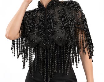 May include: A black beaded capelet featuring intricate floral patterns and long, dangling fringe accents. The garment is constructed from sheer black fabric heavily embellished with black beads and sequins, worn over a solid black dress.