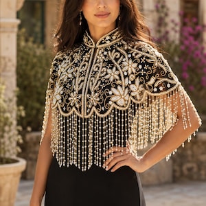 May include: A black capelet featuring an intricate high collar and elaborate gold embroidery with white floral patterns. The garment is adorned with long, dangling beaded fringe made of small white pearls that drape over the shoulders and chest.