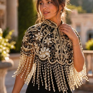 May include: A black capelet featuring intricate gold embroidery and white floral beadwork. The garment has a high collar and a scalloped hemline adorned with long, dangling strands of white pearls and clear beads that drape over the shoulders.