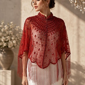 May include: A deep red sheer capelet featuring intricate beadwork, teardrop-shaped red crystals, and a high collar. The garment has a scalloped hemline adorned with long, delicate red fringe that hangs down over a plain white sleeveless dress.
