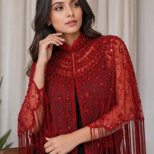 May include: A deep red sheer capelet featuring intricate beadwork, teardrop-shaped crystals, and a high collar. The garment is adorned with dense embroidery and long, delicate beaded fringe along the hem, creating a textured, layered appearance.