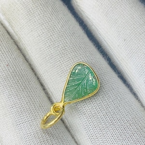 May include: A gold pendant featuring a leaf-shaped green gemstone. The pendant has a detailed leaf pattern carved into the green stone and is set in a gold frame. The pendant is approximately 2.5 cm in length.