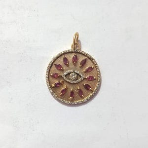 May include: Gold-tone pendant featuring an eye design. The pendant has a central eye shape with a diamond-like center and is surrounded by red gemstone accents. The outer edge is lined with small, clear stones.