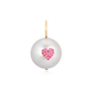 May include: A large, round, white pearl pendant with a small heart-shaped design made of pink gemstones. The pendant has a gold-colored loop at the top for hanging.