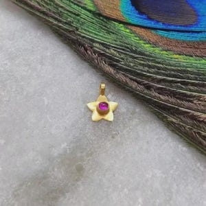 May include: A gold-toned star-shaped pendant featuring a small, circular, vibrant pink gemstone set in the centre. The pendant has a smooth, matte finish and includes a small loop at the top for attachment to a chain or cord for jewellery.
