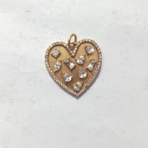 May include: A gold-colored heart-shaped pendant with a textured surface. The pendant is encrusted with small, clear gemstones around the edges and larger, irregularly shaped gemstones in the center. A small loop is at the top for a chain.
