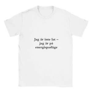 Puede incluir: Camiseta blanca con texto negro "Jag är inte lat - jag är på energisparläge". El texto se traduce como "No soy vago, estoy en modo ahorro de energía". La camiseta tiene cuello redondo y mangas cortas.