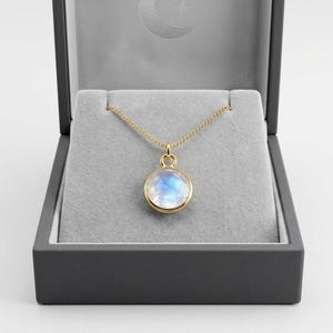 Op de afbeelding: Een goudkleurige ketting met een ronde, iriserende hanger, gepresenteerd in een grijze sieradendoos. De hanger heeft een gouden rand en ketting. De doos is donkergrijs met een grijs interieur.