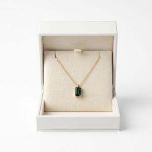 Puede incluir: Un collar dorado con un colgante rectangular de piedra preciosa verde esmeralda se muestra en una caja de joyería blanca abierta. El collar se presenta sobre un interior color crema. La cadena es fina y delicada.